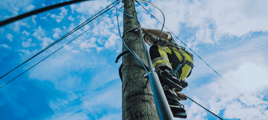 OneCo worker working up on a telephone pole, photo taken from below with blue sky and clouds visible in the background