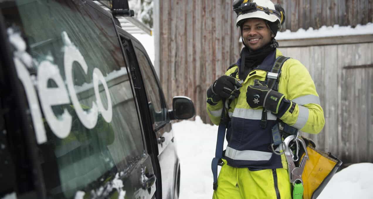 A OneCo worker is seen smiling at the camera whilst standing next to an open car door. They are dressed in climbing gear.