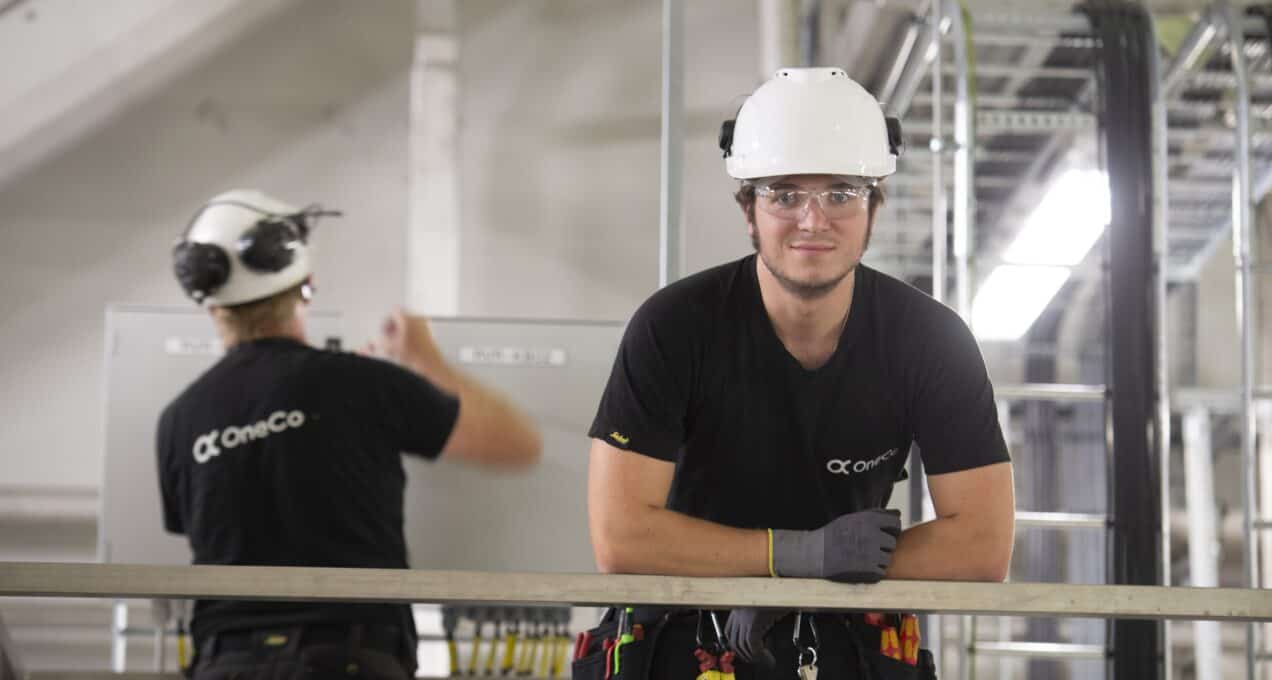 A OneCo worker is looking directly at the camera with helmet and glasses on. In the background another worker is seen fixing an electric outlet.