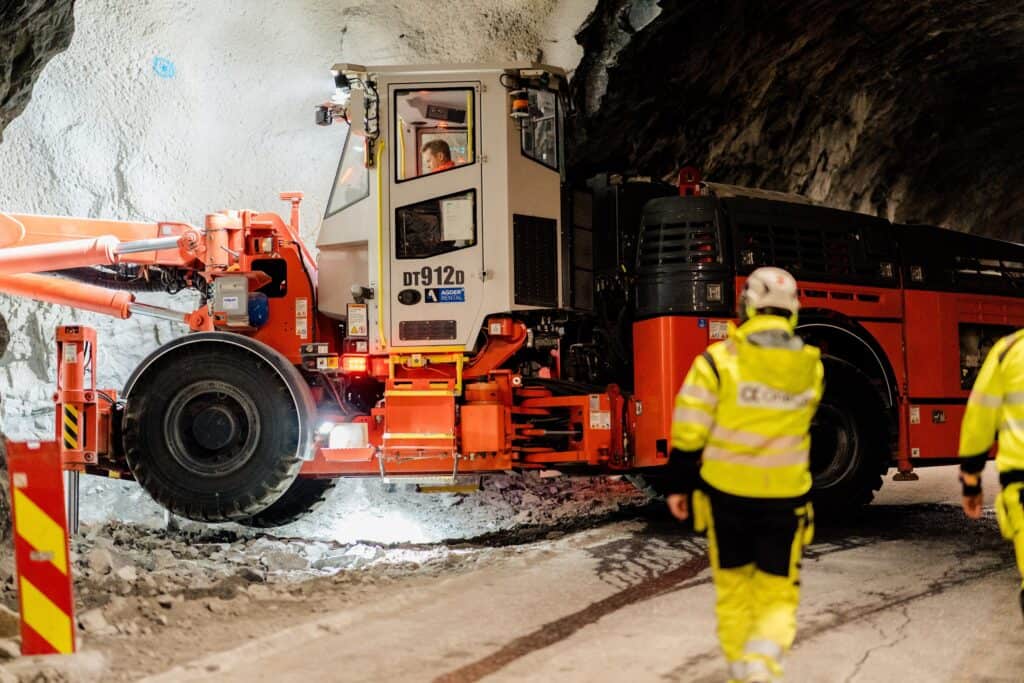 A drilling machine is pictured digging out the side of a tunnel. The back of two workers are visible in the foreground.