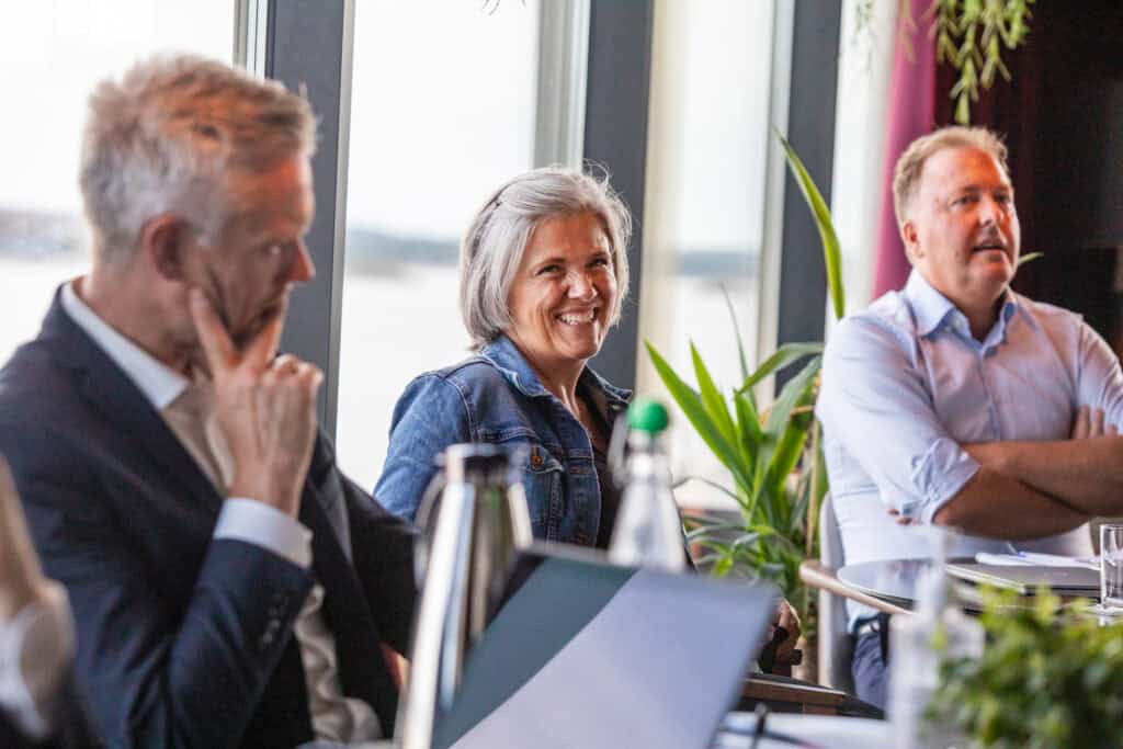 Three people are visible sitting around a table in a meeting, the one on the middle is smiling at the camera.