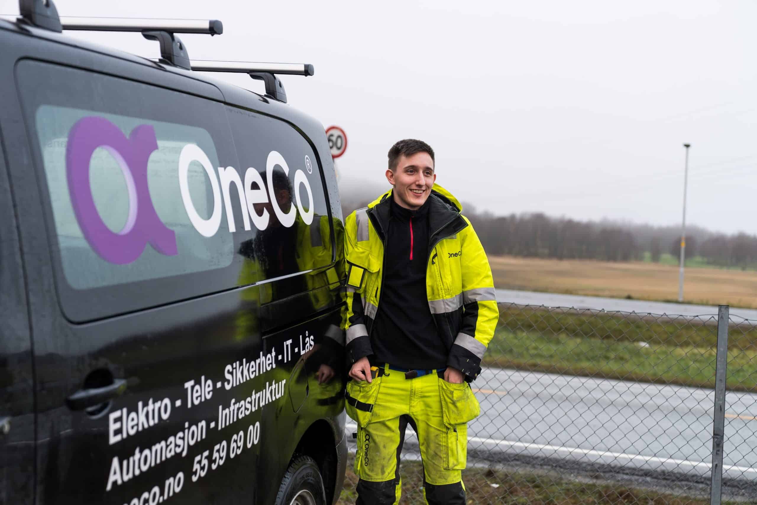 OneCo lærlinger leaning against his work car with hands in pockets smiling into the distance