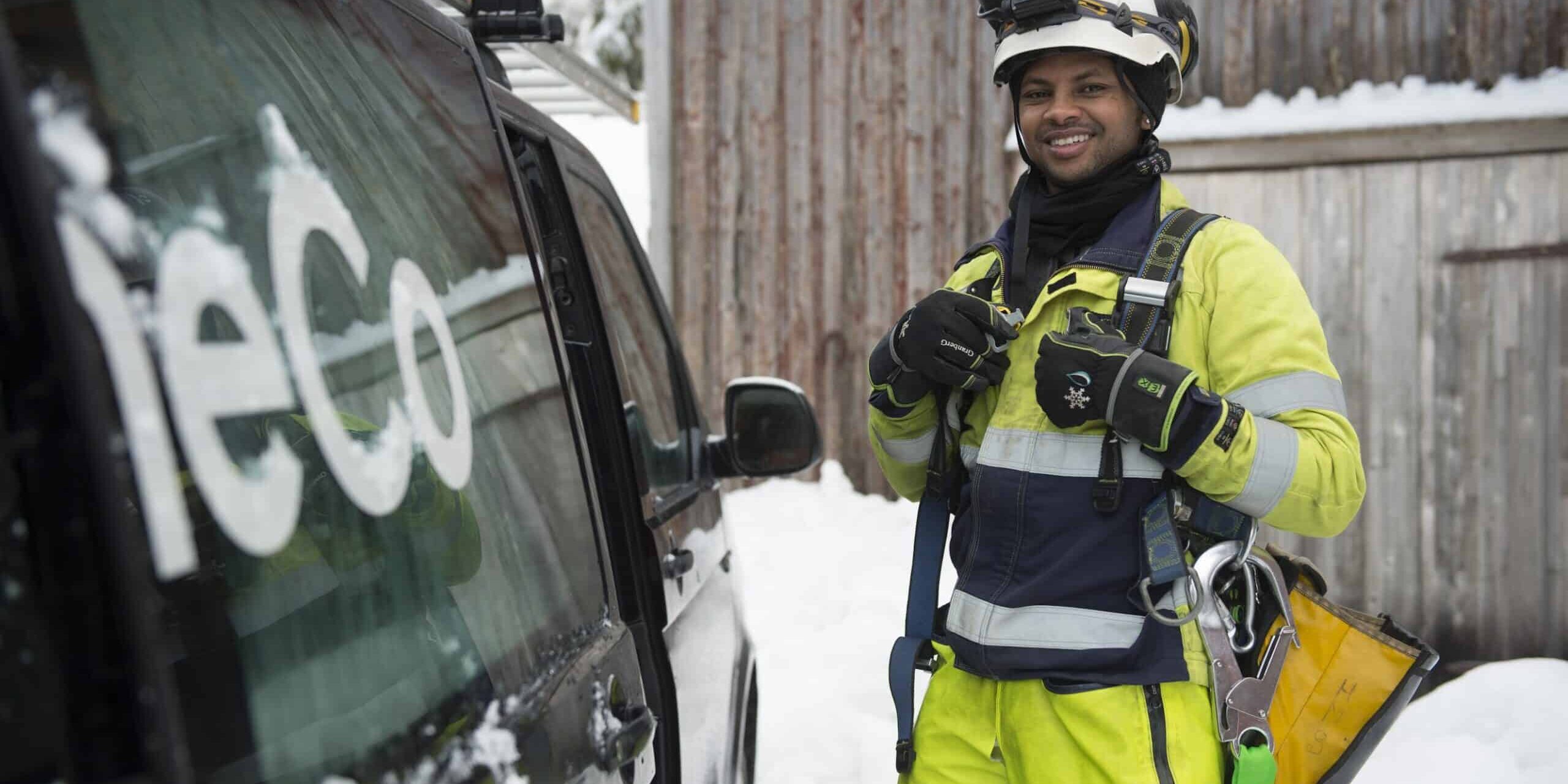A OneCo worker is seen smiling at the camera whilst standing next to an open car door. They are dressed in climbing gear.