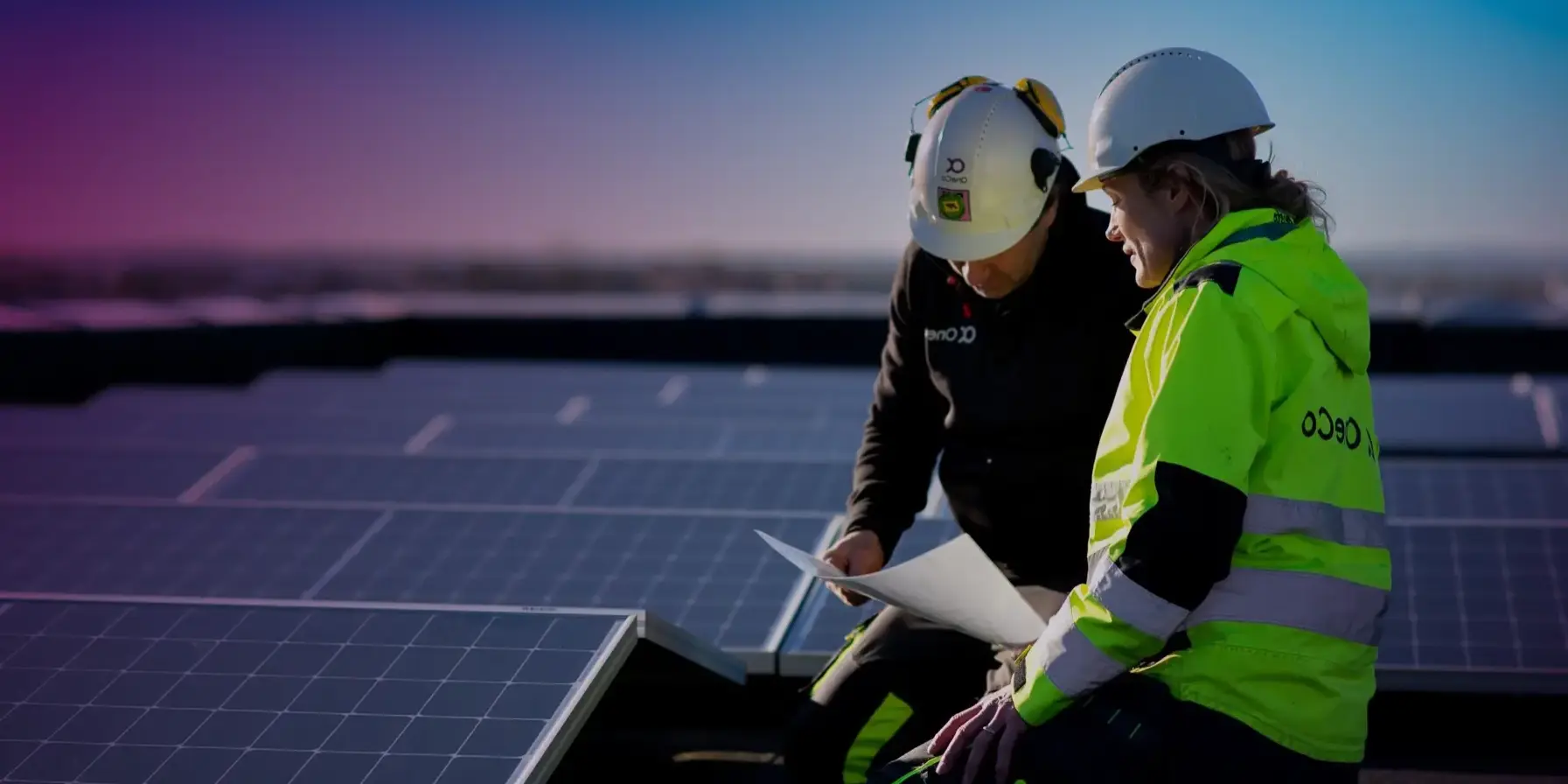 Two OneCo workers look together at a paper whilst seated next to a large solar system installation