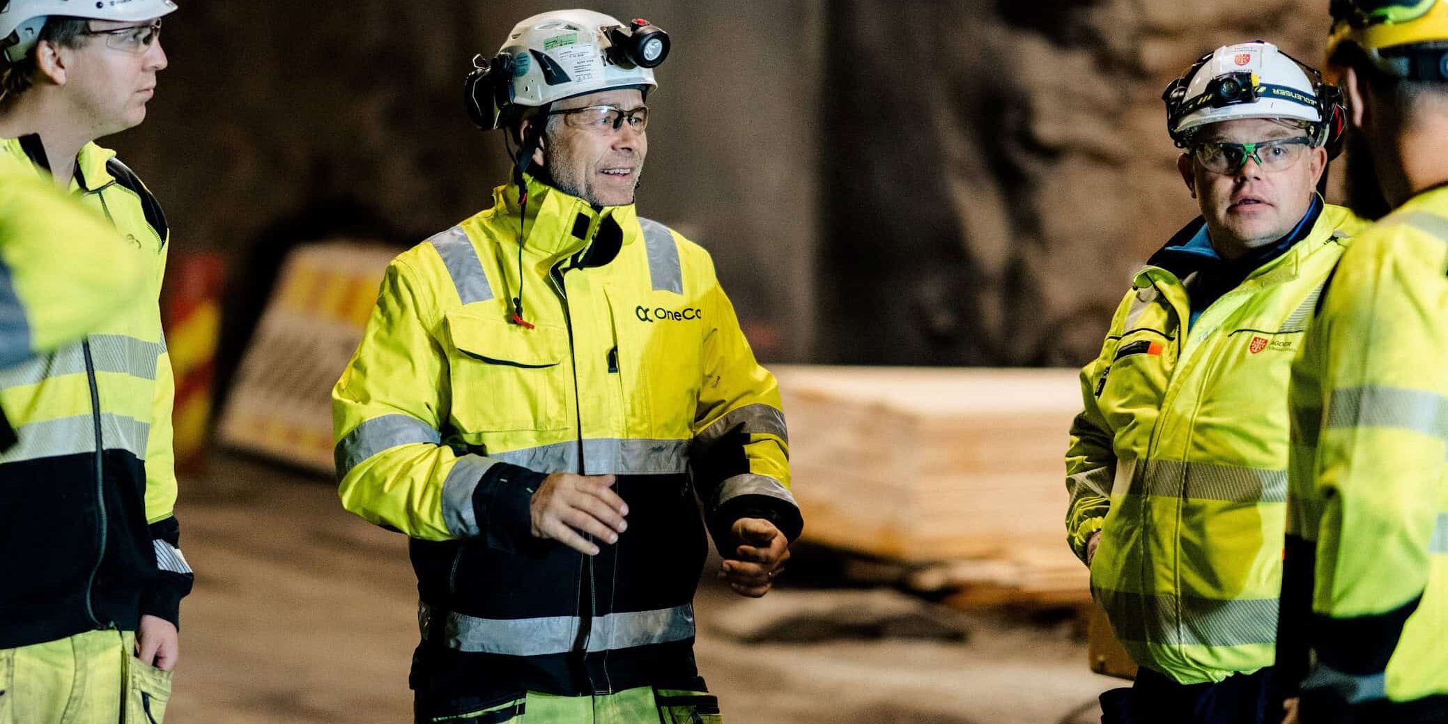 A group of OneCo workers are seen meeting together on the job with helmets and uniforms on.